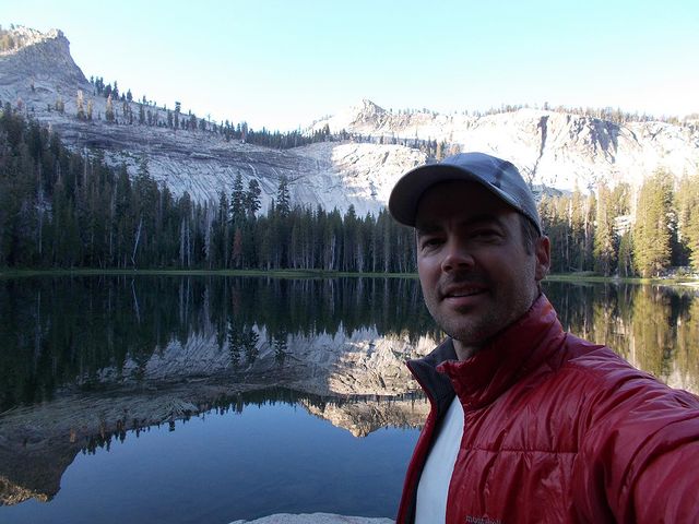 Matt Long in a red jacket and grey cap in front of a lake and hill.